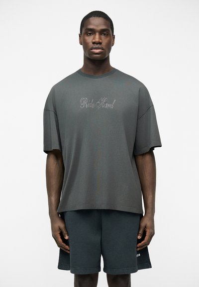 Homme portant un T-shirt ample gris foncé avec le texte "Ride Hard" en cursive et un short assorti gris foncé, sur un fond blanc uni.