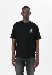 Black cotton t-shirt with short sleeves and a round neckline. Features a small graphic on the left chest. Worn with gray jeans.