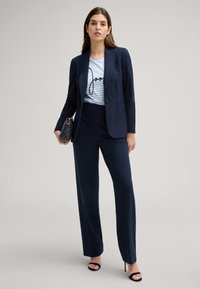 Blazer su misura blu navy sopra una t-shirt a righe con un design grafico nero. Pantaloni a gamba larga blu navy e sandali con tacco neri. Borsa nera.
