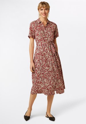 Czerwona sukienka w kwiaty z krótkimi rękawami, kołnierzykiem i wiązaniem w talii. Materiał zdobią białe wzory liści, a sukienka ma rozkloszowany dół. Do kompletu czarne baleriny.