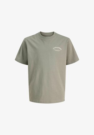 Garçon debout et souriant, portant un t-shirt vert clair Jack & Jones, un jean bleu clair et des chaussures beiges, sur un fond uni.