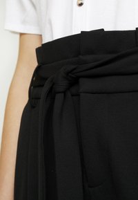 Svarta högmidjade byxor med ett brett tygbälte knutet framtill. Materialet verkar mjukt, med veck och sidfickor.