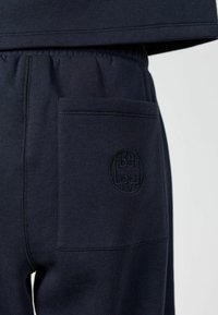 Tumed sinised dressipüksid, millel on tagataskul õmmeldud ümmargune logo, kantud sobiva tumeda sinise pealsega.