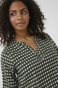 Young woman with curly hair wearing a green and white diamond-patterned V-neck blouse, looking to her left against a plain background.