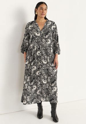 Lange zwart-wit bloemenjurk met een V-hals, driekwartmouwen en een gelaagde rok, gecombineerd met zwarte enkellaarsjes.