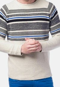 Personne portant un pull en tricot crème avec des rayures horizontales noires, grises et bleues, mains jointes, et pantalon bleu sur un fond blanc.