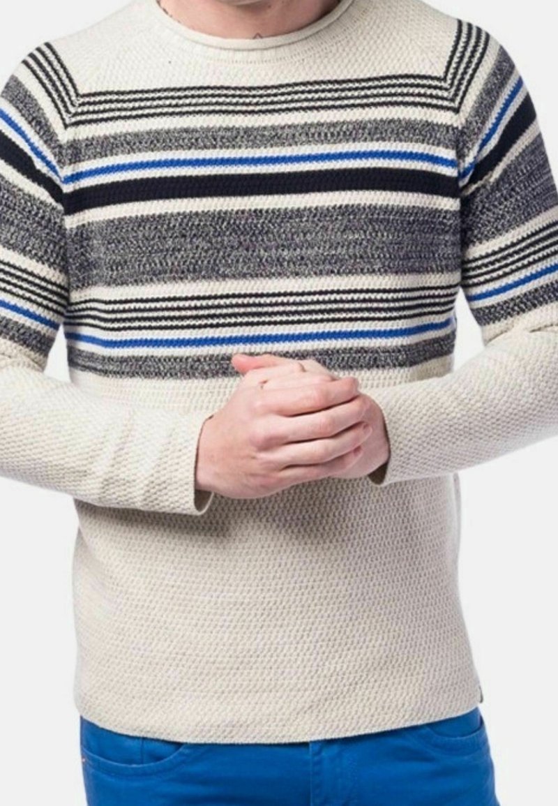 Personne portant un pull en tricot crème avec des rayures horizontales noires, grises et bleues, mains jointes, et pantalon bleu sur un fond blanc.