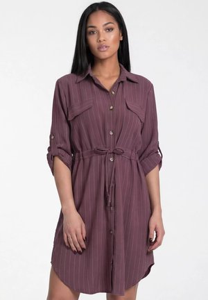 Kobieta ubrana w mauve sukienkę w cienkie prążki zapinaną na guziki, z podwiniętymi rękawami i przewiązanym paskiem w talii, stojąca na jednolitym jasnym tle.