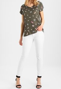 Frau trägt ein olivgrünes kurzärmliges Oberteil mit Schmetterlings- und Insektenmotiven, weiße, taillierte Hose und schwarze, offene Sandalen mit Absatz, steht mit einer Hand in der Tasche.