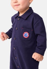 Marineblå bomulls jumpsuit med lange ermer, knappeåpning og en brystlomme med et rundt logopatch med røde detaljer.
