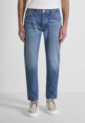 Man die lichtblauwe jeans met rechte pijpen, witte sneakers met dikke zolen en een wit shirt draagt, staand tegen een effen achtergrond.