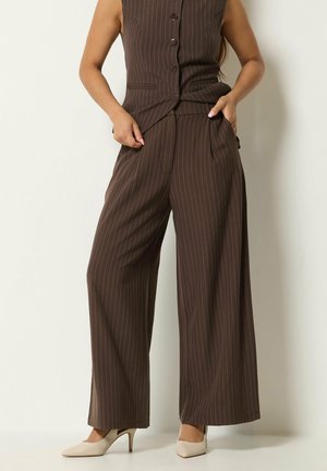 Femme portant un pantalon large à fines rayures marron et un gilet assorti, avec des chaussures à talons beiges, debout sur un fond clair.
