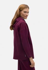 Sudadera con capucha burdeos hecha de tela suave, con un corte relajado, hombros caídos y puños de canalé. Diseño minimalista, sin logotipos visibles.