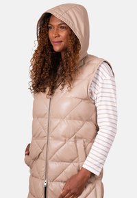 Woman with curly hair wearing a beige quilted sleeveless hooded vest over a white and beige striped long-sleeve shirt, hands in pockets.