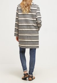 Lungo cappotto beige con strisce geometriche nere, caratterizzato da una linea dritta e tasche laterali, indossato sopra jeans blu e sandali marroni.