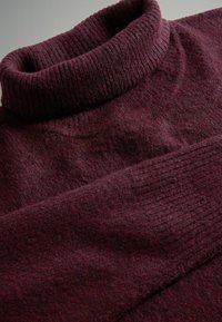 Detailní záběr na složený bordó vlněný rolák se žebrovaným límcem a manžetami, zobrazující měkkou texturu látky.