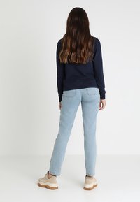 Navy long-sleeve shirt, light blue straight-leg jeans, beige sneakers with textured soles. Back view shows long, wavy hair.