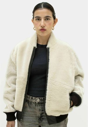 Kvinde med mørkt hår iført en hvid sherpa-jakke, sort ribbet top og grå jeans, stående mod en ensfarvet lys baggrund.