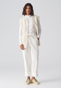 Gilet texturé de couleur claire sur un haut à manches longues blanc, associé à un pantalon blanc à côtes et des chaussures beiges à lacets avec des semelles compensées.