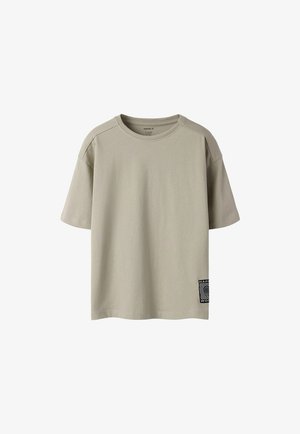 Lyse beige t-shirt med korte ærmer og rund hals med et lille sort-hvidt mærke nær den nederste kant på venstre side.