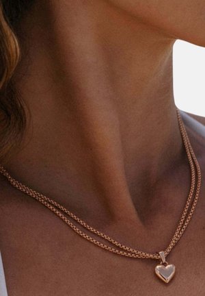Nahaufnahme eines Halses mit einer doppelten goldenen Halskette und einem herzförmigen Anhänger vor einem einfarbigen Hintergrund.