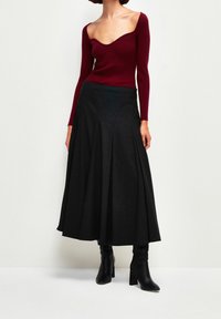 Burgundy ribbed pitkähihaine paita, jossa on avara kaula-aukko, yhdistettynä mustiin raitapolkimiin; mallilla mustat korkeat saappaat.