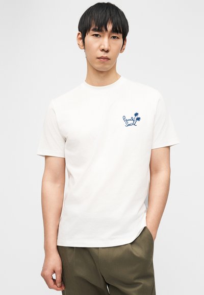 Άνδρας που φοράει λευκό μπλουζάκι με μικρό μπλε φοίνικα και λογότυπο κειμένου στο αριστερό στήθος και χακί παντελόνι, στέκεται μπροστά σε απλό φόντο.