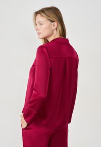 Bordowa satynowa bluzka z długimi rękawami, spiczastym kołnierzykiem, kieszonką na piersi i detalem z zakładką z tyłu. Gładka faktura i dopasowany krój.