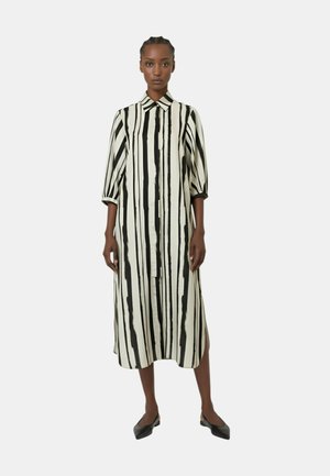 Donna in piedi, che indossa un vestito di media lunghezza a righe verticali bianche e nere, con bottoni, maniche al gomito e ballerine nere con punta.