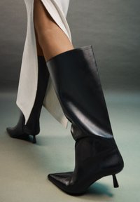 Bottes en cuir noir montantes avec un bout pointu et un talon aiguille, présentant une finition lisse et un design ajusté.