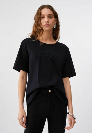 Frau mit welligem Haar, die ein schwarzes T-Shirt mit dem Aufdruck "Good Morning", schwarze Hosen, ein silbernes Armband und große silberne Ohrringe trägt, steht vor weißem Hintergrund.