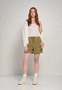 Pantaloni corti verde oliva con vita regolabile, abbinati a una canotta bianca e a un cardigan leggero e fluido. Stivali da combattimento beige con suole spesse.