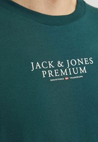 Teal crew-neck puuvillainen t-paita, jossa valkoiset tekstit "JACK & JONES PREMIUM" ja pieni rekisteröity tavaramerkki symboli alla. Pehmeä tekstuuri.