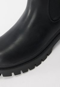 Botín negro de goma hasta el tobillo con una textura suave, que cuenta con paneles laterales elásticos y una suela robusta con tacos para mayor agarre.