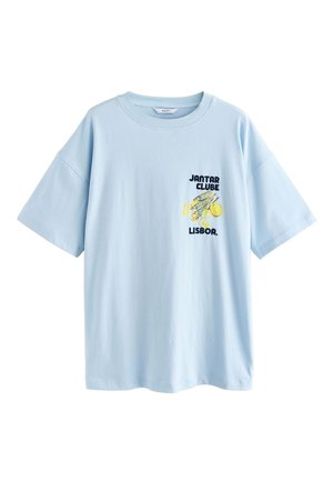 Femme aux cheveux bouclés portant un T-shirt blanc oversize et un bas de bikini fleuri, debout près d'une falaise en bord de mer avec une bouteille sur un mur en pierre.