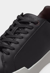 Melna ādas sneaker ar melnām šņorēm, uz mēles iespiests logotips un balta gumijas zole ar šūtu detaļu.