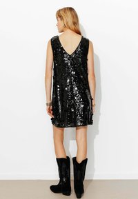 Robe noire à sequins avec un dos en V profond et un design sans manches, associée à des bottes de cowboy noires montantes. La robe présente une surface brillante et texturée.