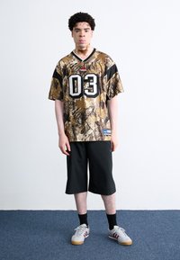 Camo-mønstrede sportstræk i nuancer af brun og grøn med sorte accenter, der har et stort '03' på fronten. Sammen med sorte shorts.