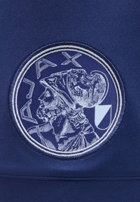 Στρογγυλή επιφάνεια με λεπτομερές προφίλ ενός ανδρός με γένια που φορά κράνος και τη λέξη "AJAX" από πάνω σε σκούρο μπλε ύφασμα.