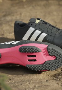 Schwarze Adidas-Radschuhe mit weißen Streifen, rosa und schwarzen Klettsohlen, auf dem Erdreich platziert.