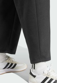 Pantalons de jogging noirs avec une coupe droite et un ourlet côtelé, associés à des baskets blanches avec des rayures noires et des chaussettes de sport blanches avec des rayures noires.