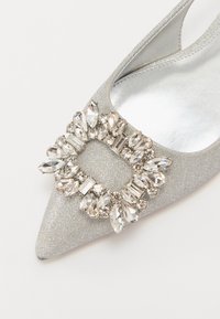 Escarpin argenté à talons hauts avec une pointe fine, texture scintillante, et une boucle en cristal décorative ornée de pierres transparentes de différentes formes.