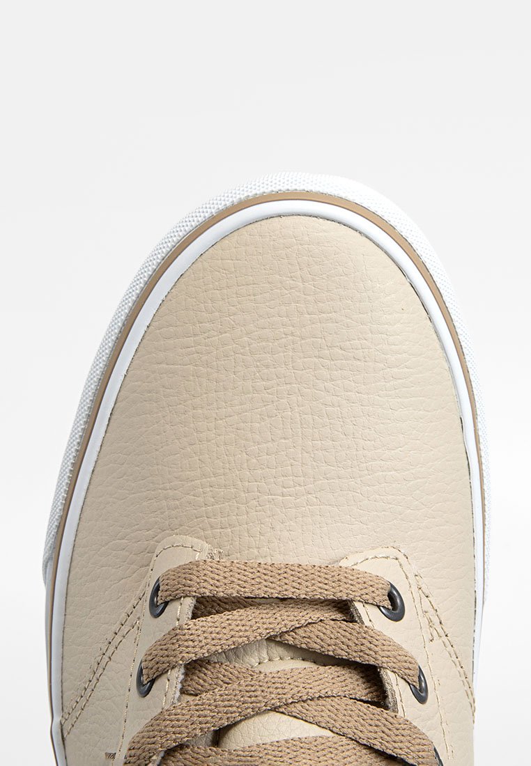 Acercamiento de la punta de una zapatilla de cuero beige con cordones texturizados y suela blanca con borde marrón sobre un fondo blanco.