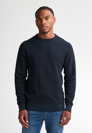 Man with short curly hair wearing a dark textured sweater and blue jeans standing against a plain light background.