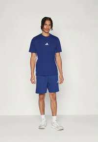 Μπλε αθλητική μπλούζα και σορτς σετ, με λευκές τρεις ρίγες και λογότυπο. Το υλικό φαίνεται ελαφρύ. Τα παπούτσια είναι ανοιχτού γκρι.