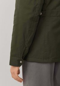 Chaqueta verde oliva con textura suave, que presenta un puño con cierre de presión, costuras sutiles y un corte recto, mostrada desde el lateral con una mano.