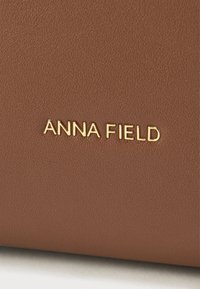 Barna texturált bőr aranyozott "ANNA FIELD" logóval, sima felülettel és minimalista dizájnnal.