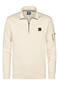 Maglione color crema con colletto alto e chiusura a mezza zip. Presenta un patch quadrato nero e una tasca con zip sulla manica. Materiale in maglia morbido.