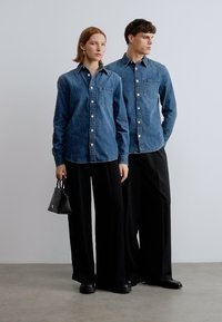 Dve osebi, oblečeni v jeans srajce z gumbi, črne hlače z širokimi hlačnicami in črne čevlje. Ena drži majhno črno torbico. Svetlo ozadje.