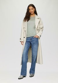 Langer cremefarbener Mantel mit schwarzen Knöpfen, grauer strukturierten Pullover und ausgestellte blaue Jeans. Silberne spitz zulaufende Schuhe runden das Outfit ab.